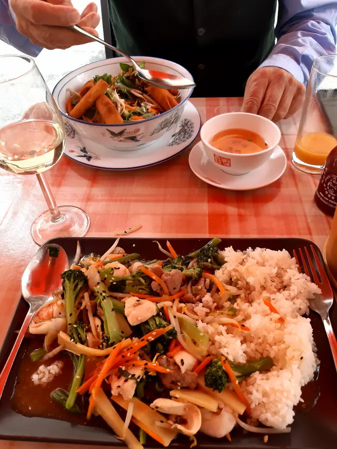 Gedeckter Tisch mit kariertem Tischtuch: Wok-Gericht mit Garnelen, Brokkoli und Reis, dahinter eine Schale mit Sommerrollen