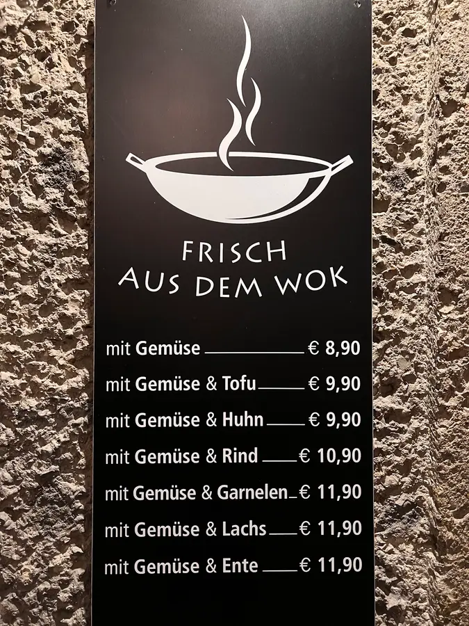 Schwarzes Schild mit Wok-Symbol an der Steinmauer: Frisch aus dem Wok, mit Gemüse, Tofu, Huhn, Rind, Garnelen, Lachs oder Ente